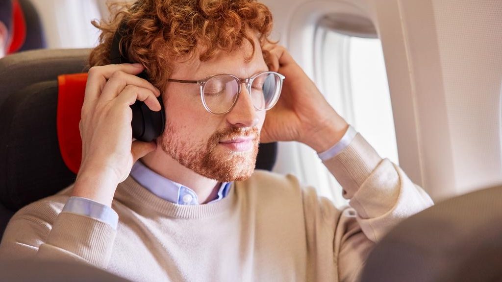 A man with curly red hair and glasses wearing headphones rests in an airplane seat with eyes closed.
