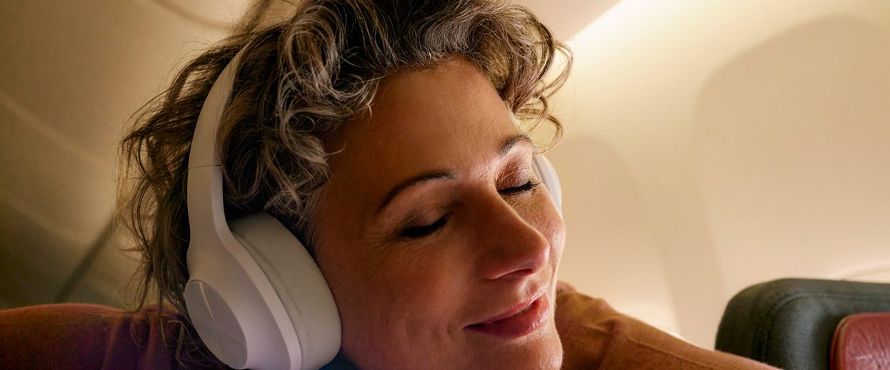 Nahaufnahme einer lächelnden Frau mit geschlossenen Augen, die im Flugzeug Kopfhörer trägt.