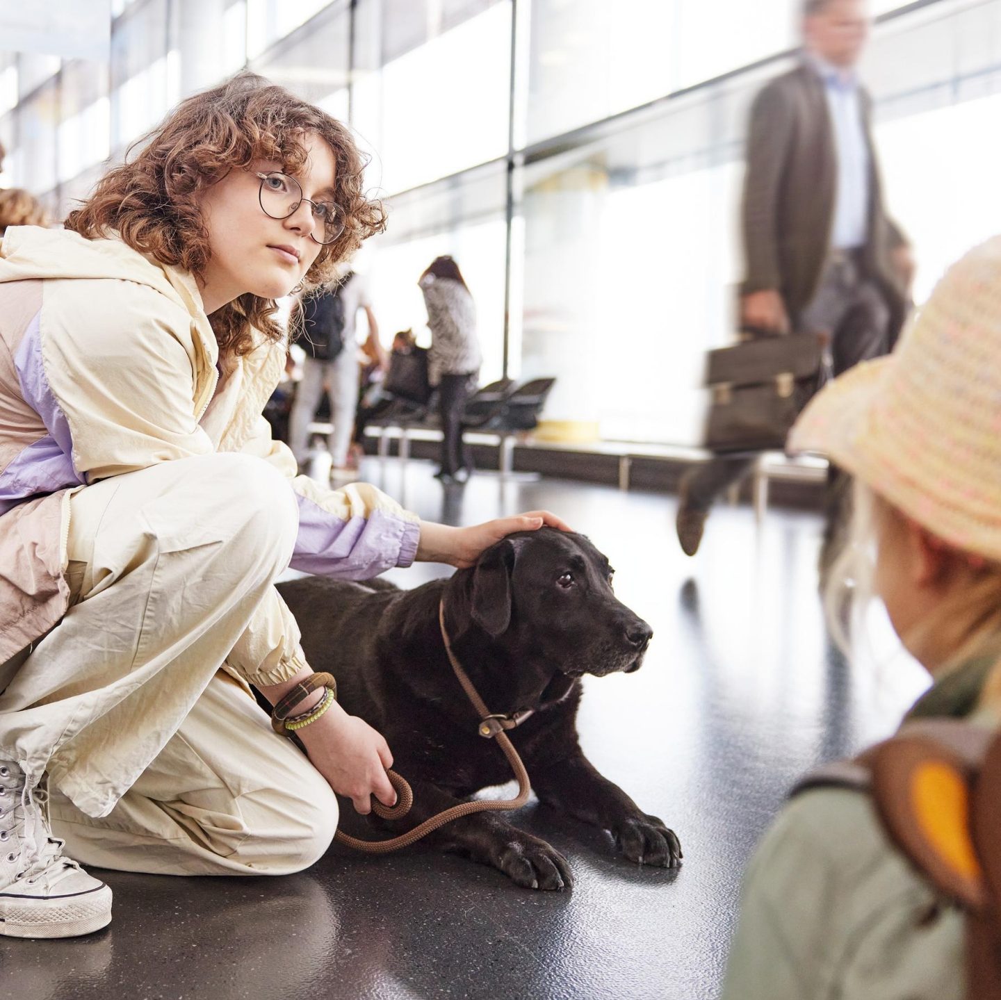 Person mit Hund sitzt in einem Flughafenterminal, während Reisende vorbeigehen. Im Vordergrund steht ein Kind mit Rucksack und betrachtet den Hund.