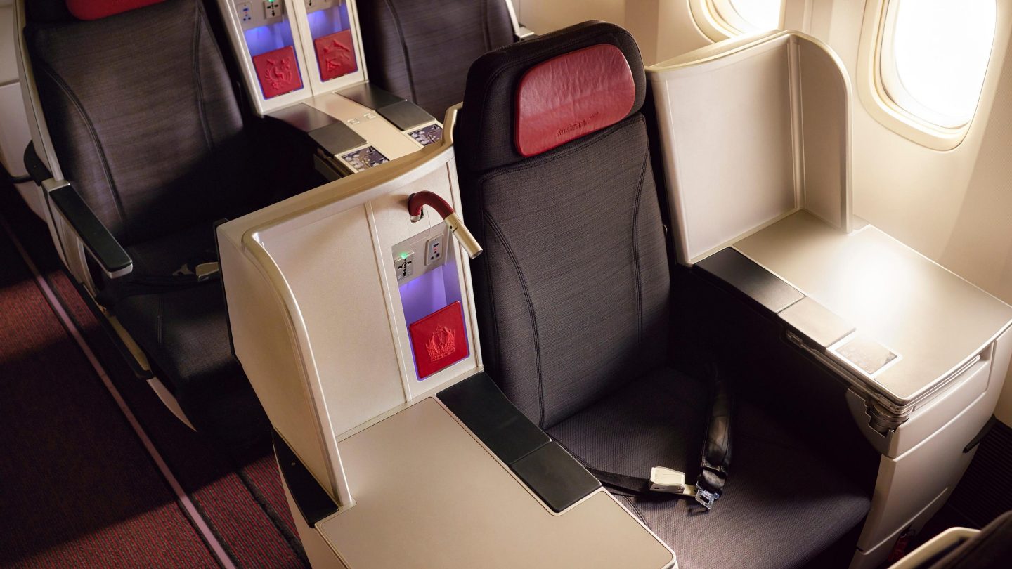 Business class airplane seats with red and black upholstery, separated by a console with storage and power outlets.