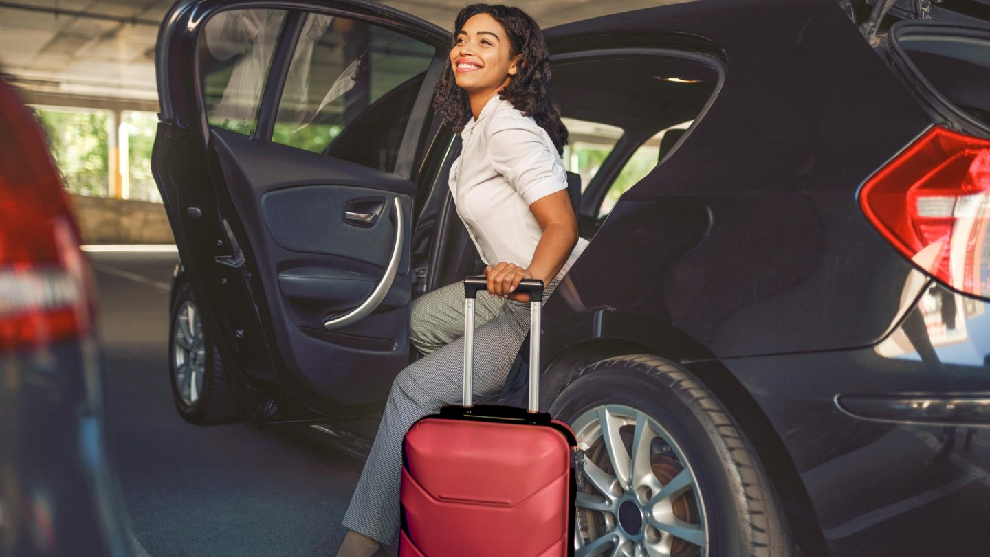 Happy woman with suitcase gets into the car on parking. Female traveler with luggage in vehicle park lot, passenger with bag. Girl with baggage near automobile