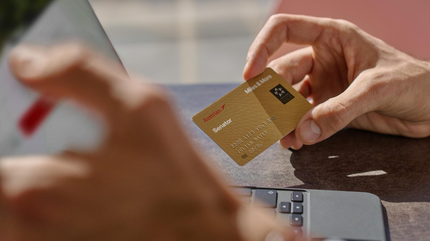 A gold credit card is held by a person in front of a laptop.