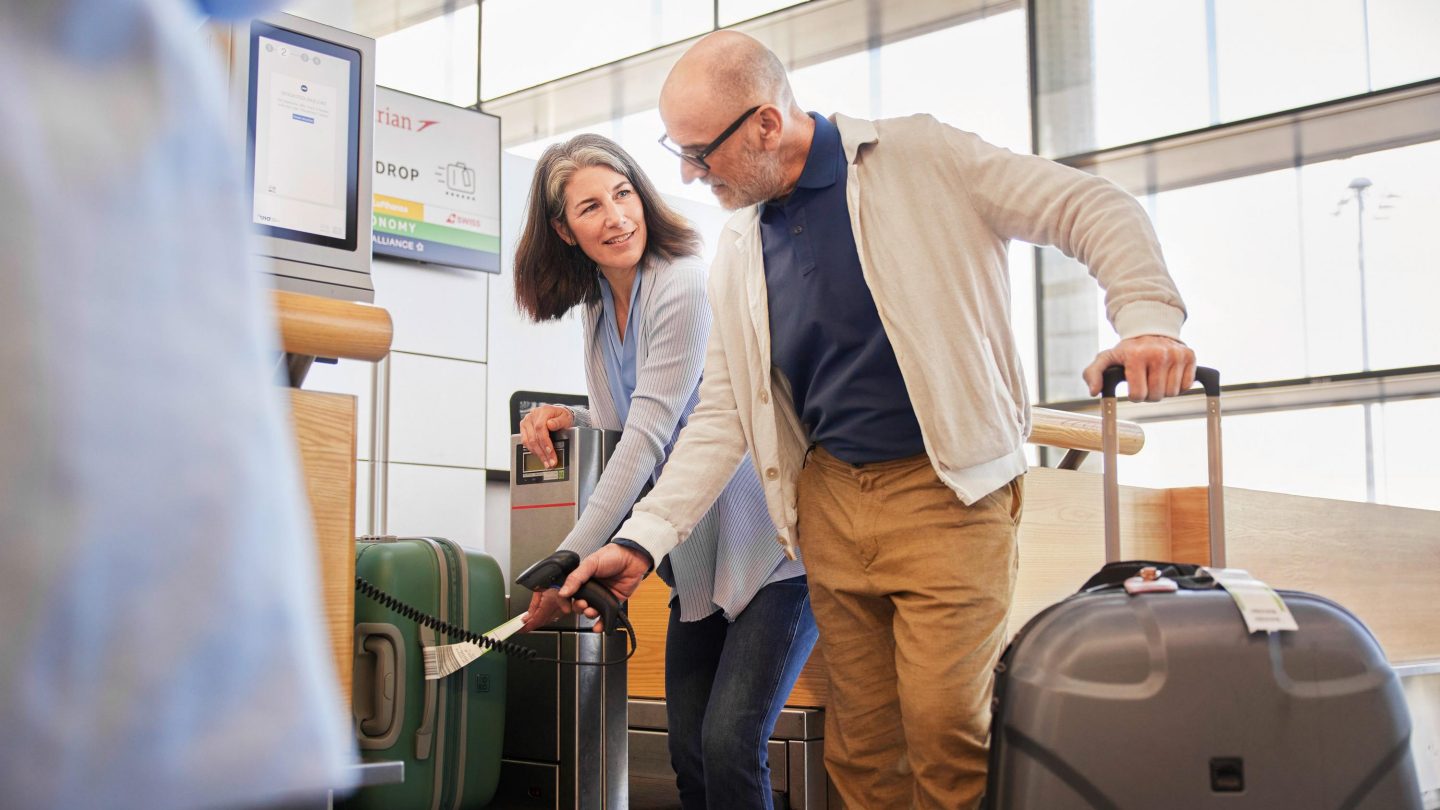 Two travelers placing suitcases at an airport baggage check-in kiosk.