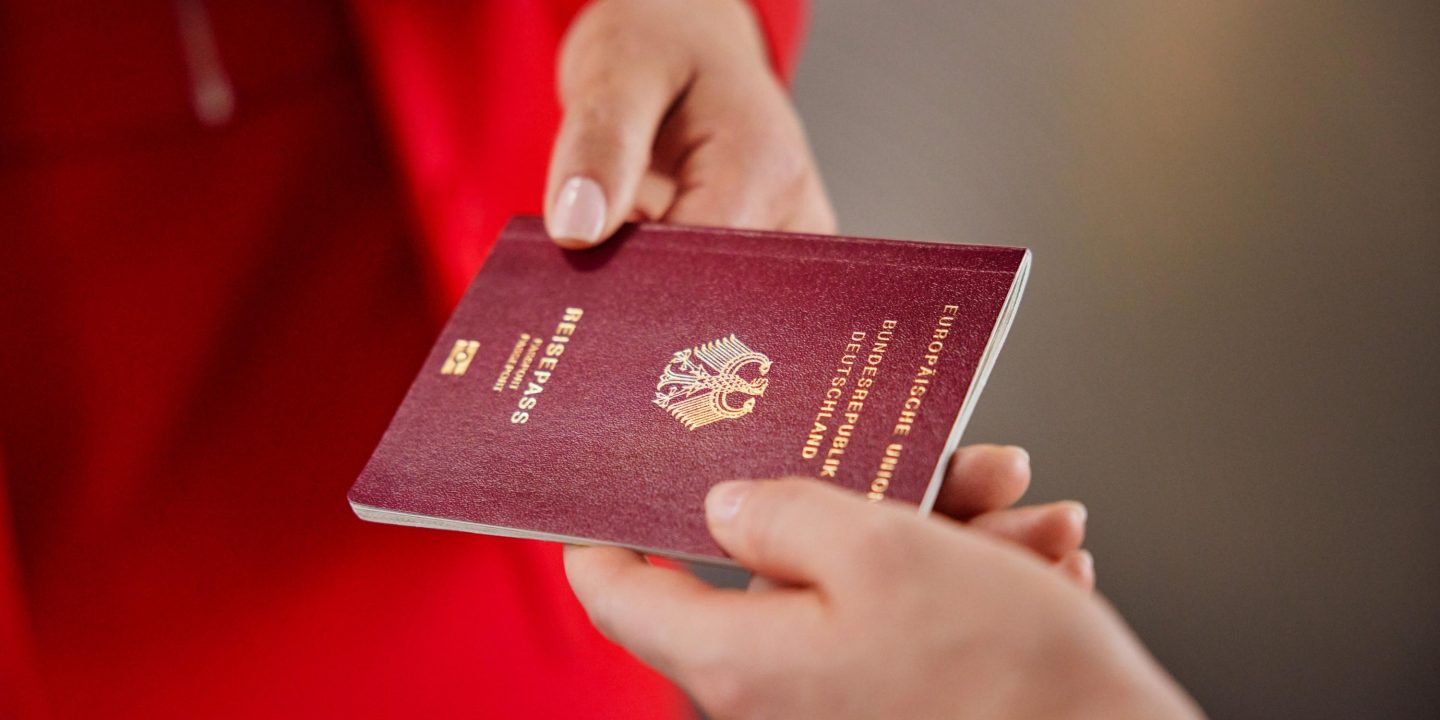 Eine Hand überreicht einen burgunderroten deutschen Reisepass an eine andere Person.