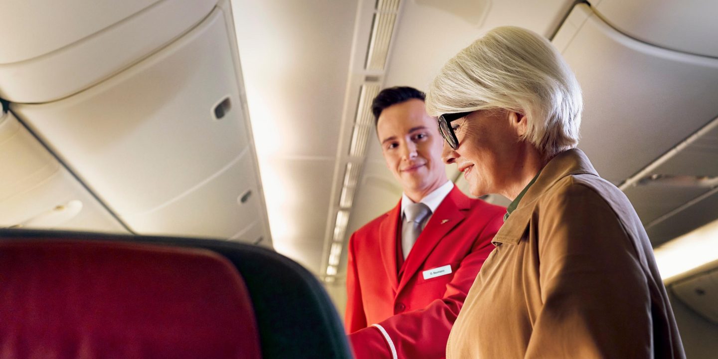 A flight attendant in a red uniform helps a female passenger to her seat on an airplane.
