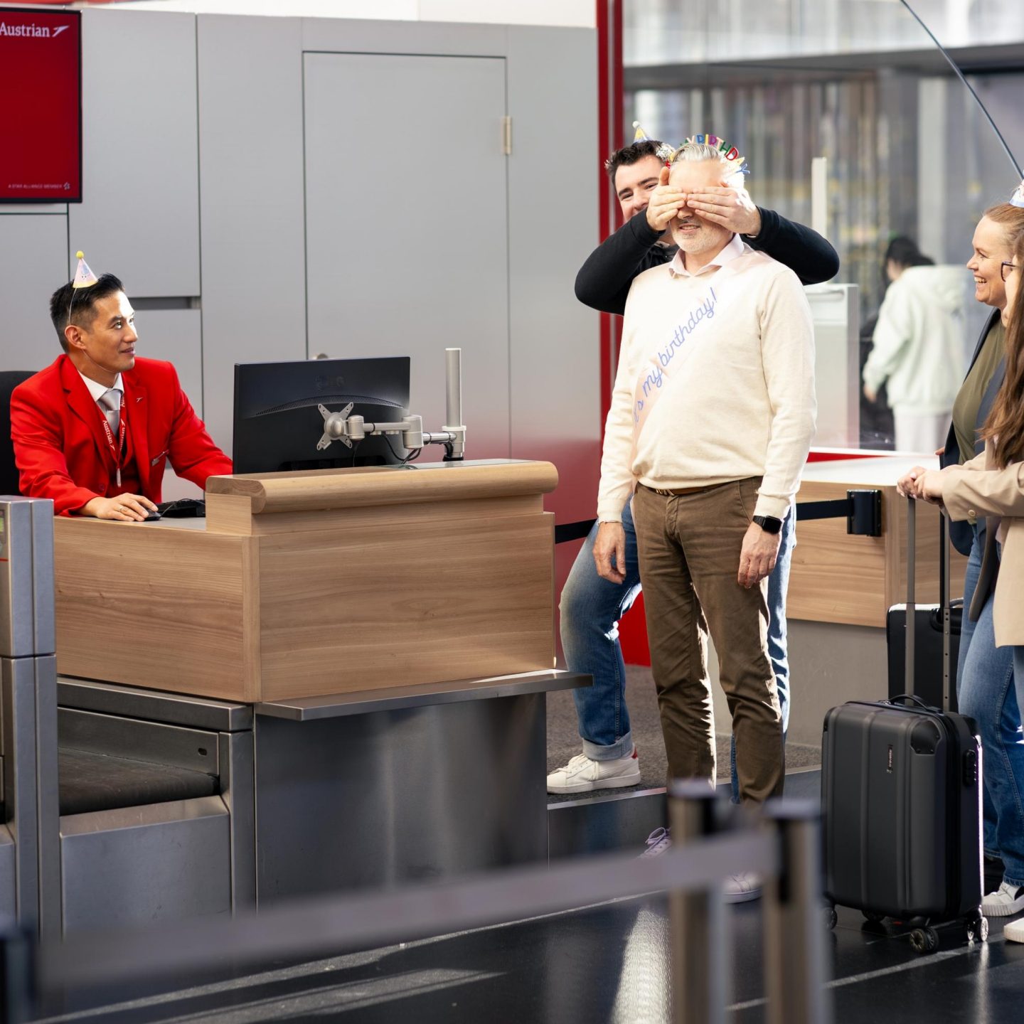 Check-in-Szene am Flughafen: Ein Mitarbeiter in roter Uniform sitzt hinter dem Schalter, während ein Mann mit Partyhut davor steht und von zwei Personen überrascht wird. 