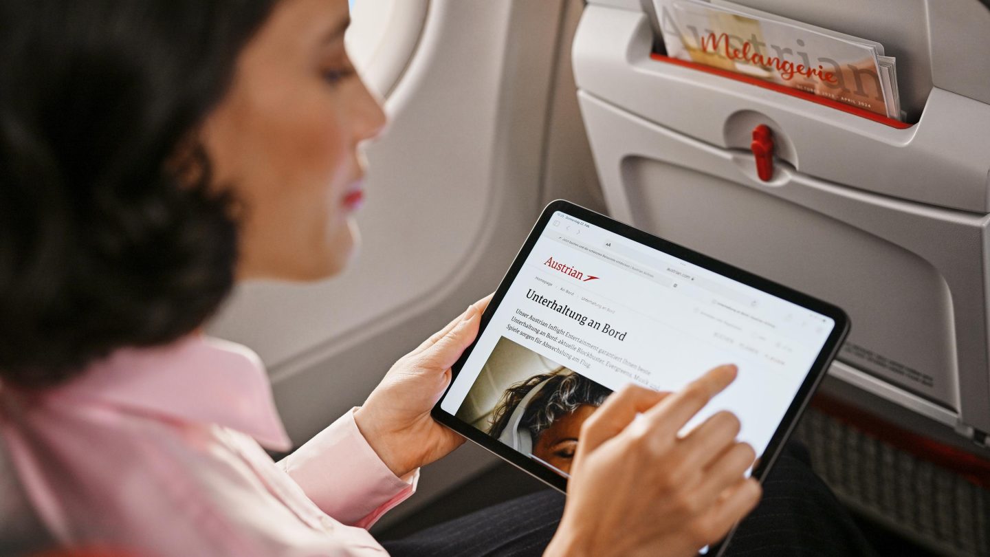 A person using a tablet on an airplane, reading an article from Austrian Airlines about in-flight entertainment.