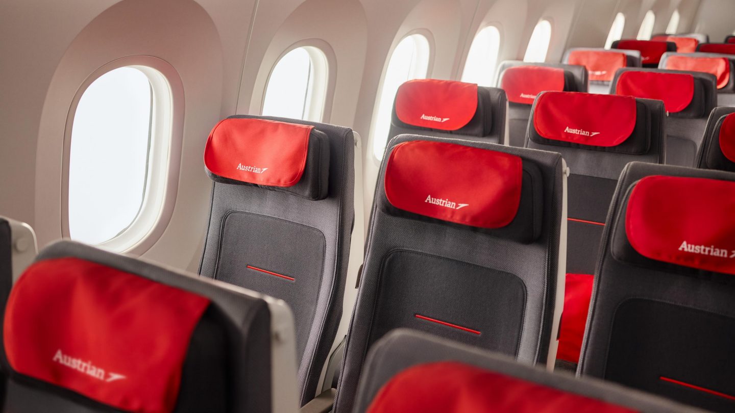Airplane cabin interior with rows of gray seats and red headrests featuring the Austrian Airlines logo.