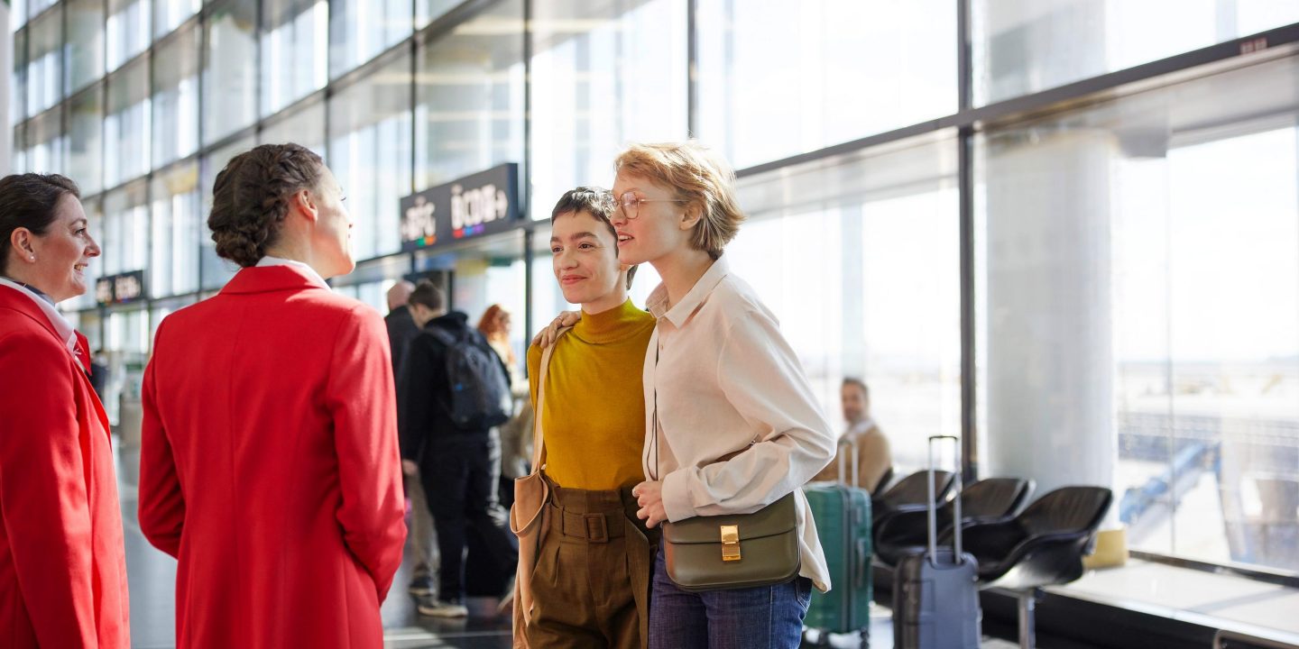 Lächelndes Flugpersonal begrüßt zwei zufriedene Passagiere in einem hellen Flughafenterminal.