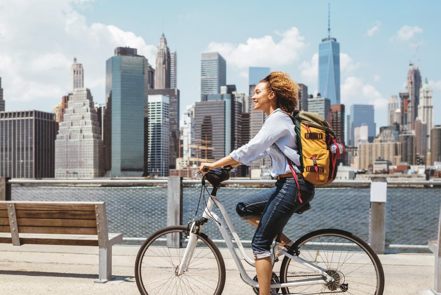 Eine Frau fährt mit dem Fahrrad entlang der Hafenpromenade. Im Hintergrund erhebt sich die markante Skyline von New York mit ihren Wolkenkratzern vor dem Himmel. Die Szene vermittelt urbanes Flair und eine entspannte Atmosphäre am Wasser.