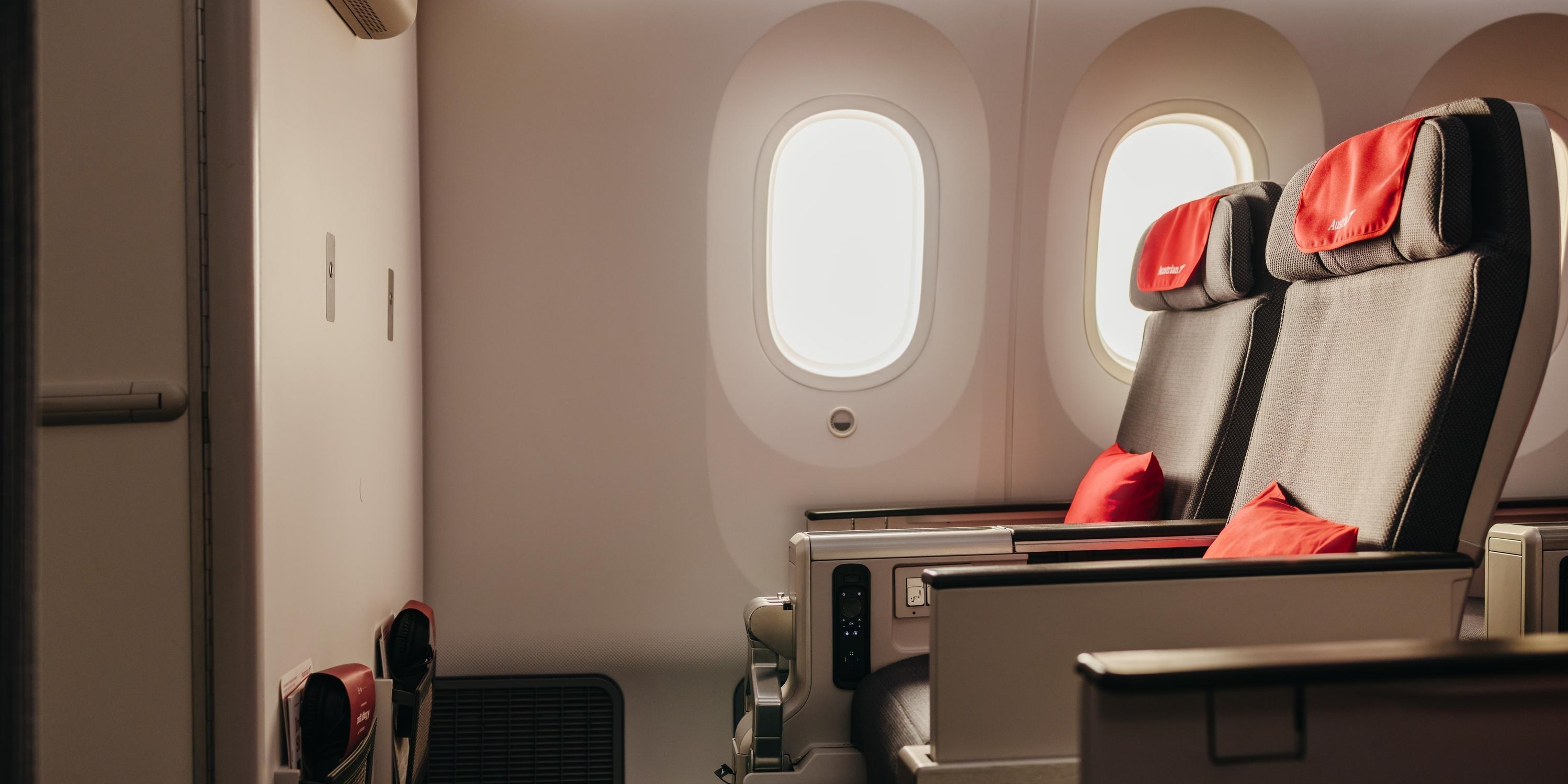 Spacious airplane cabin with two wide seats, red headrests and pillows, softly lit by two oval windows.