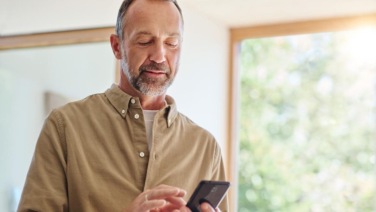 Man in a brown shirt using a smartphone indoors near a window with blurred greenery outside.