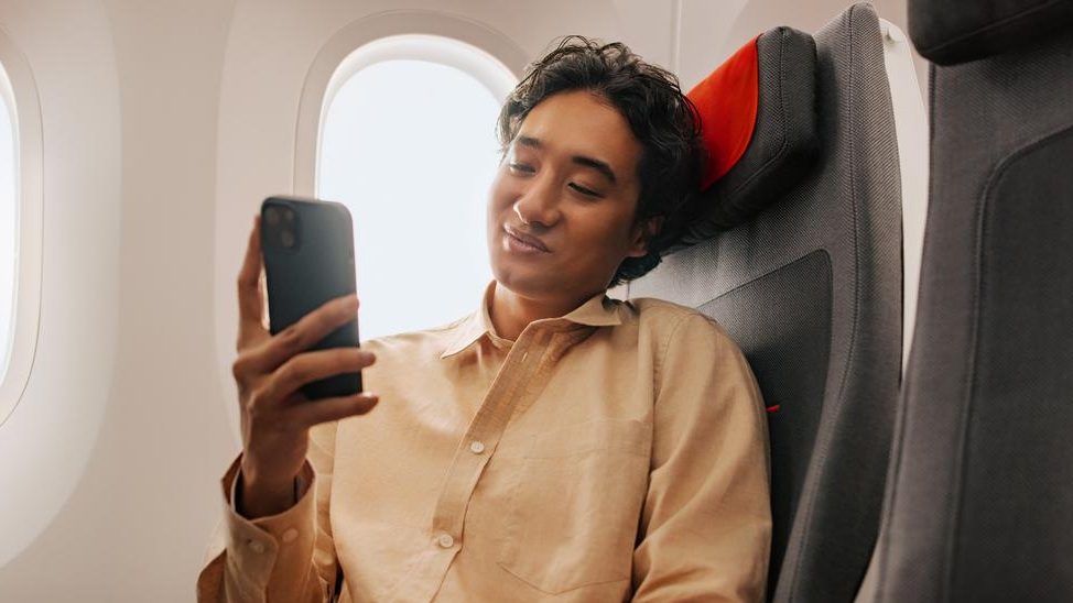 Passenger in beige shirt using smartphone while seated next to airplane window during flight.