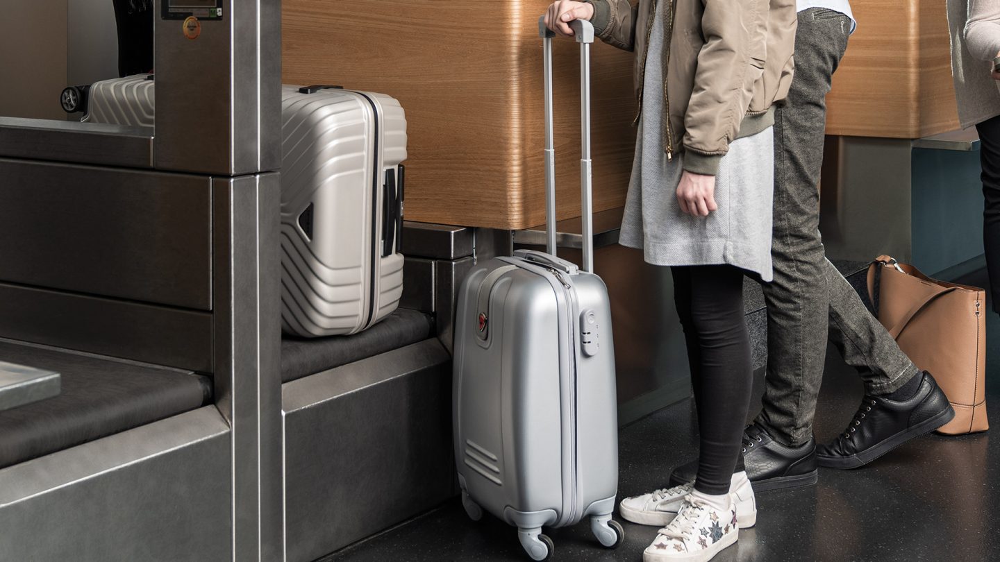 Traveler at check-in with a suitcase; another on the conveyor belt.
